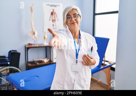 Mittelalterliche Frau mit grauen Haaren, die in der Schmerzheilungsklinik arbeitet, lächelt fröhlich und bietet Handfläche als Hilfestellung und Akzeptanz an. Stockfoto