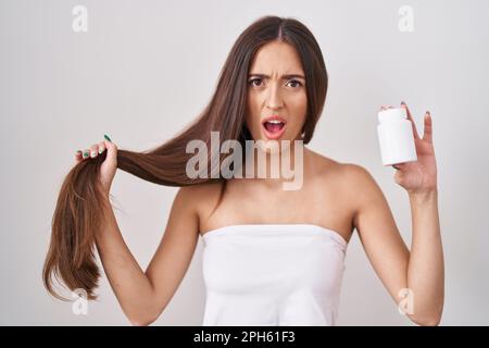 Junge hispanische Frau, die Haare mit der Hand und Pillen im Schockgesicht hielt, skeptisch und sarkastisch aussah, überrascht von offenem Mund Stockfoto