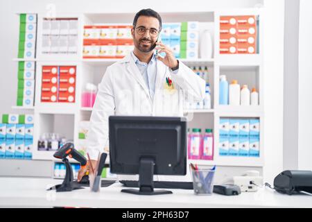 Junger hispanic-Mann Apotheker im Gespräch auf Smartphone mit Computer in der Apotheke Stockfoto