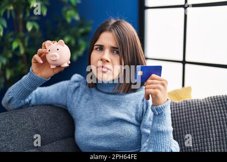 Junge hispanische Frau mit Sparschwein und Kreditkarte, ahnungslos und verwirrter Ausdruck. Zweifelhaftes Konzept. Stockfoto