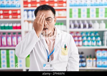 Chinesischer junger Mann, der in einer Apotheke arbeitet und müde gähnt, indem er Gesicht, Augen und Mund mit der Hand bedeckt. Das Gesicht tut weh. Stockfoto