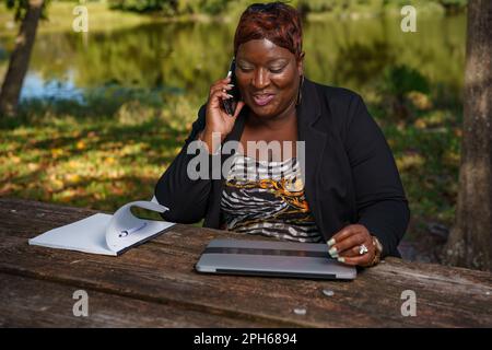 Erfolgreiche schwarze Geschäftsfrau, die geschäftlich telefoniert, während sie im Park arbeitet Stockfoto