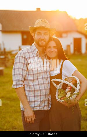 Porträt eines jungen glücklichen Bauernpaares, das bei Sonnenuntergang Korb mit frischem Bio-Gemüse auf seinem rancho hält. Vegetarisch gesunde Lebensmittel. Familie Stockfoto