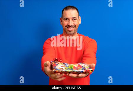 Bärtiger hispanischer Mann in seinen 40s Jahren trägt einen roten Pullover, der seiner Geliebten ein Geburtstagsgeschenk überbringt. Er lächelt mit Zufriedenheit, wie es sein Partner getan hat Stockfoto