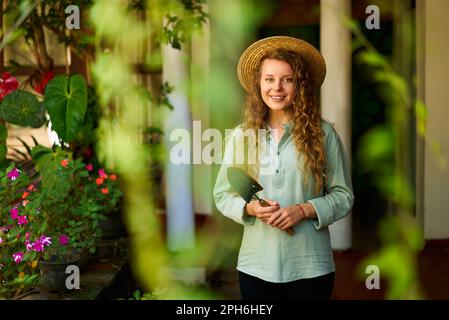 Eine junge Frau steht im Garten und hält Gartenwerkzeug in der Hand. Sie lächelt und schaut in die Kamera. Junge Gärtnerin mit einer Kelle in Grün Stockfoto