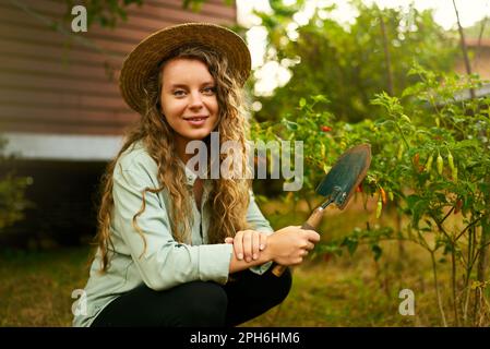 Junge Gärtnerin lächelt auf ihrer Farm, die zwischen Pflanzen sitzt. Glückliche Weiße in ihrem Garten mit einer Schaufel nahe der reifen Gemüseernte Stockfoto