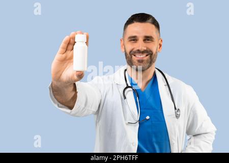 Glücklicher Mann mittleren Alters Arzt im Mantel, zeigt ein Glas mit Medikamenten isoliert auf blauem Hintergrund, selektiver Fokus, Nahaufnahme Stockfoto