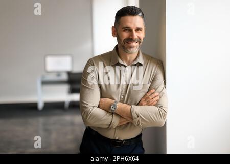 Porträt eines selbstbewussten Geschäftsmannes mittleren Alters, der im Bürosaal steht, mit gekreuzten Händen posiert und sich an die Wand lehnt Stockfoto