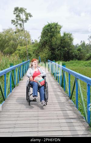 Reife hispanische Frau, die im Rollstuhl sitzt, sich mitten auf einer Brücke posiert, während sie an einem Park im Freien spaziert. Stockfoto