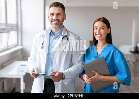 Porträt von Mann und Frau des Doktors, die in der Klinik posieren und vor der Kamera lächeln, Klemmbretter halten, Labormäntel tragen Stockfoto