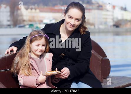 Eine Mutter und Tochter entspannen sich auf einer Bank am Ufer der Moldau und trinken dabei den auf dem lokalen Bauernmarkt gekauften Toast Stockfoto