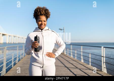 Sportliches, Schwarzes Weibchen Mit Einer Wasserflasche In Den Händen, Die Draußen Am Pier Steht Stockfoto