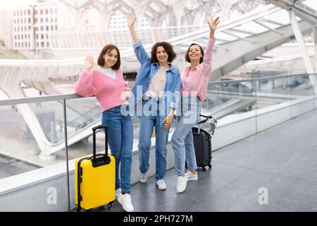 Drei Fröhliche Weibliche Freunde, Die Sich Vor Der Kamera Die Hände Winken, Während Sie Am Flughafen Posieren Stockfoto