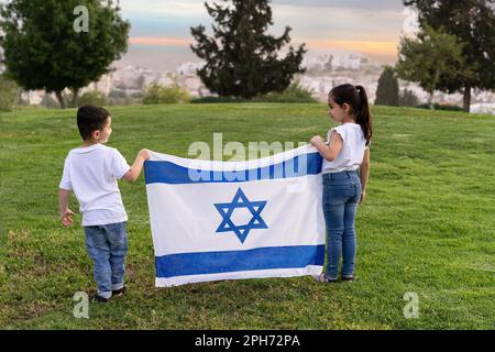 Kleine Kinder stehen auf einem Hügel und halten eine israelische Flagge. Ein kleines Mädchen und ein Junge mit einer israelischen Flagge im Sonnenuntergang. Stockfoto