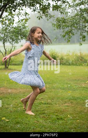 Fröhliches, süßes kleines Mädchen in einem Sonnenkleid, tanzend und auf grünem Rasen unter dem Regen. Barfußkind genießt den Sommer im Freien. Stockfoto