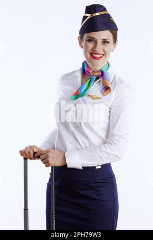 Lächelnde, stilvolle Stewardess vor weißem Hintergrund in Uniform mit Rolltasche. Stockfoto