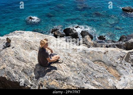 Ein Mann mit Hut sitzt auf einem Stein nahe dem türkisfarbenen Meer, Griechenland, Kreta Stockfoto