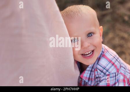 Nahaufnahme eines kleinen Jungen im karierten Hemd, umarmtes Mütterbein. Draufsicht auf ein lächelndes Kleinkind mit blauen Augen. Der Begriff des elterlichen Schutzes, t Stockfoto