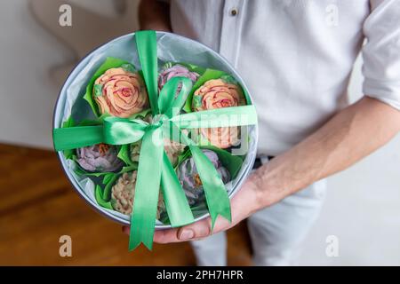 Ein gesichtsloser Mann hält eine runde Schachtel mit süßen Marshmallow-Blumen in den Händen. Geschenk mit grünem Band. Zarte, zuckerfreie Rosen. Kleine Unternehmen zu Hause. Hobby-BA Stockfoto