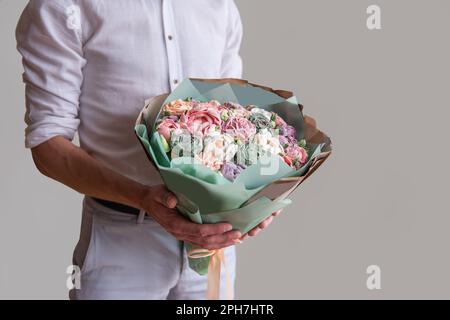 Ein gesichtsloser Mann hält einen Strauß süßer Marshmallow-Blumen in den Händen. Geschenk in pastellgrünem Umschlag mit zarten, zuckerfreien Rosen auf grauem Hintergrund. Klein Stockfoto