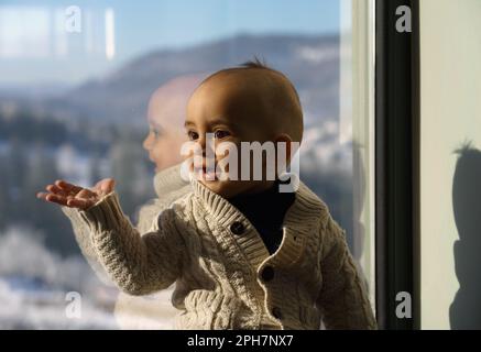 Süßer kleiner Junge, der neben einem Fenster sitzt und einen Pullover trägt Stockfoto