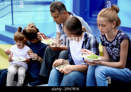 Gruppe hungriger Menschen, die Essen mit Farben der ukrainischen Flagge im Hintergrund essen. Hilfe für Flüchtlinge im Krieg Stockfoto