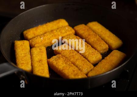 Panierte Fischstäbchen in einer Bratpfanne. Herstellung von gefrorenen Fischstäbchen. Fastfood. Dunkler Hintergrund. Stockfoto