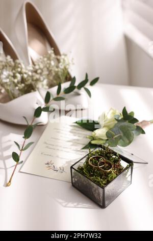 Wunderschöne Eheringe in Glaskiste, Blüten, Brautschuhe und Einladung auf weißem Tisch drinnen Stockfoto