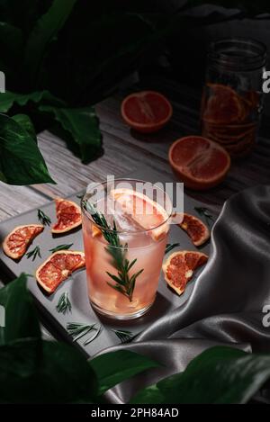 Bartisch mit Grapefruit-Cocktail, Run, Gin und Whiskey, garniert mit getrockneten Früchten und Rosmarin, im Hintergrund ein Glasgefäß, Pflanzen und Grapefrui Stockfoto