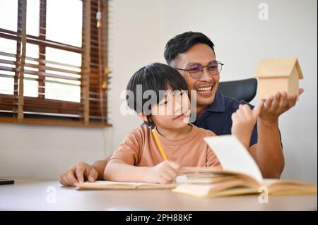 Der freundliche und fröhliche asiatische Vater bringt seinem kleinen Sohn bei, Hausaufgaben zu machen oder ein Hausmodell auf Papier zu zeichnen. Ein glückliches Familienzeitkonzept Stockfoto