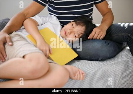 Der entspannte und glückliche junge asiatische Sohn hörte sich ein Märchen an und schlief auf dem Schoß seines Vaters auf dem Sofa im Wohnzimmer ein. Stockfoto