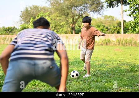 Fröhlicher, junger asiatischer Junge, der mit seinem Vater im Garten Fußball spielt, Spaß zusammen hat. Ein glückliches Familienkonzept Stockfoto