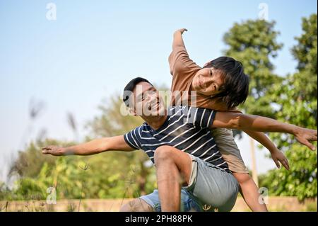 Lächelnder und glücklicher asiatischer Vater, der mit seinem Sohn im Garten spielt, während er ihn auf dem Rücken trägt. Aufgeregter und fröhlicher junger asiatischer Junge, der sich auf die Beine stellt Stockfoto