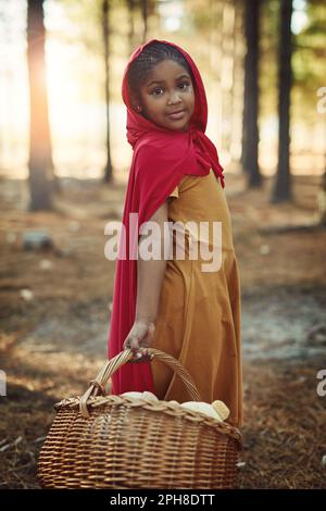 Sie hat eine kleine rote Reithaube. Porträt eines kleinen Mädchens in einem roten Umhang und mit einem Korb im Wald. Stockfoto