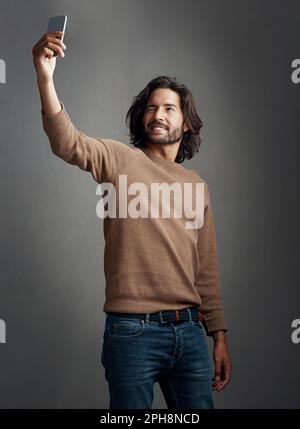 Ich zeige Ihnen das gute Aussehen. Studioaufnahme eines gutaussehenden jungen Mannes, der ein Selfie auf seinem Handy vor grauem Hintergrund macht. Stockfoto