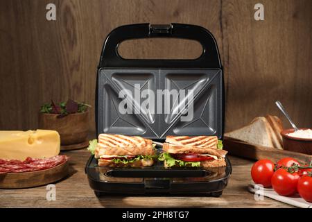 Moderne Grillmaschine mit Sandwiches und verschiedenen Produkten auf Holztisch Stockfoto