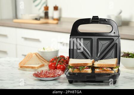 Moderne Grillmaschine mit Sandwiches und verschiedenen Produkten auf einem weißen Marmortisch in der Küche Stockfoto