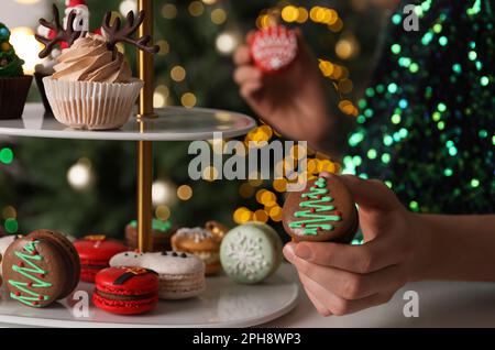 Eine Frau, die Weihnachtsmakaron aus dem Stand nimmt, gegen verschwommene festliche Lichter, Nahaufnahme Stockfoto