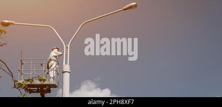 Ein Mann steht auf einem Turm und malt einen Laternenpfahl. Männer in der Wiege malen Lampenpfähle Stockfoto