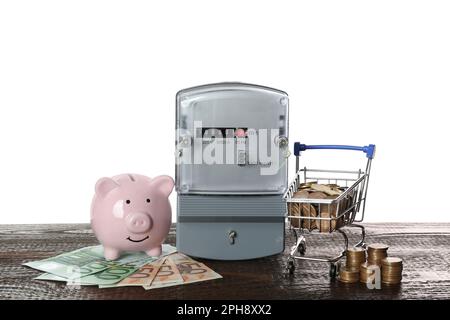 Stromzähler, Sparschwein und kleiner Einkaufswagen mit Geld auf Holztisch vor weißem Hintergrund Stockfoto