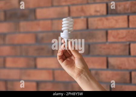 Eine Frau, die eine Leuchtstofflampe in der Nähe einer Backsteinwand hält, im Haus, Nahaufnahme. Energiesparkonzept Stockfoto