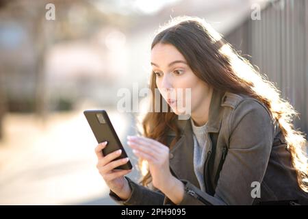 Überraschte Frau, die auf der Straße auf ihr Handy schaut Stockfoto