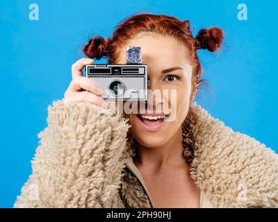 Wunderschöne Rothaarige mit einer alten 35mm-MP-Kamera und Würfelblitz auf blauem Hintergrund. Stockfoto