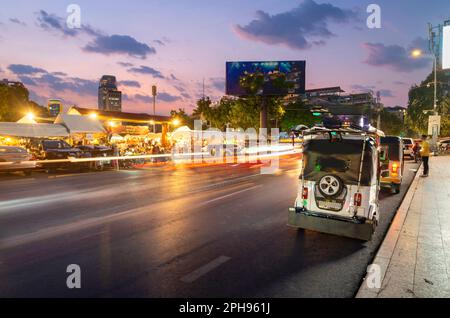 Außerhalb des geschäftigen Abendmarkts, Lichtstreifen vom vorbeifahrenden Verkehr, nach Sonnenuntergang, während Tuk Tuk Fahrer auf Passagiere warten. Die Gegend ist voller Menschen Stockfoto