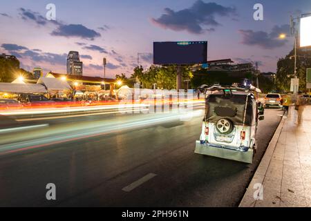 Außerhalb des geschäftigen Abendmarkts, Lichtstreifen vom vorbeifahrenden Verkehr, nach Sonnenuntergang, während Tuk Tuk Fahrer auf Passagiere warten. Die Gegend ist voller Menschen Stockfoto