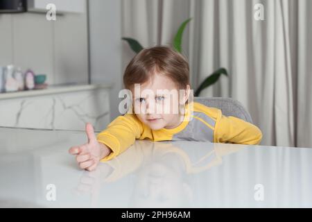 Lächelnder, glücklicher Junge in gelbem Pullover, der am Tisch sitzt und im Hintergrund der Studioküche an der Hand zieht Stockfoto