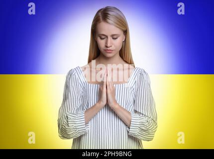 Bete für die Ukraine. Junge Frau mit gefalteten Händen betet gegen die ukrainische Nationalflagge Stockfoto