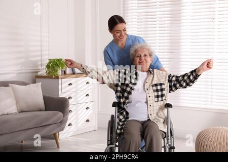 Seniorin im Rollstuhl, die körperliche Bewegung macht, und junge Pflegekraft, die ihr drinnen hilft. Häuslicher Gesundheitsdienst Stockfoto