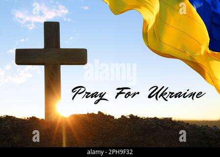 Bete für die Ukraine. Phrase, ukrainische Flagge, die bei Sonnenaufgang draußen über dem Kreuz winkt Stockfoto