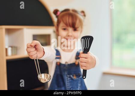 Werkzeuge in den Händen halten und es zeigen. Kleines Mädchen in lässiger Kleidung hat Spaß, wenn es mit Spielzeug in der Küche spielt Stockfoto